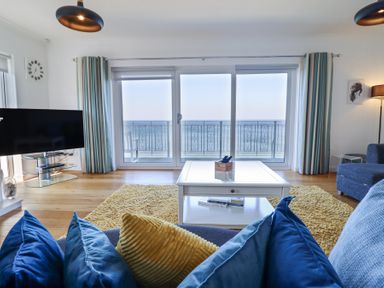 A living room with blue and yellow cushions on a sofa a white coffee table a yellow rug a television and glass doors leading to a balcony overlooking the sea at Gwylanod in Llwyngwril near Fairbourne
