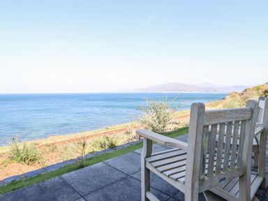 Wooden chairs on a paved patio facing the sea with railway tracks and hills in the background at Gwylanod in Llwyngwril near Fairbourne