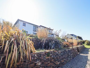 A white house behind a stone retaining wall with various plants and a gravel path at Gwylanod in Llwyngwril near Fairbourne