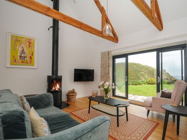 A living room with a green sofa a black coffee table a pink armchair a wood burner and large sliding glass doors showing a hill outside at Pendower Cottage in Porthgwarra
