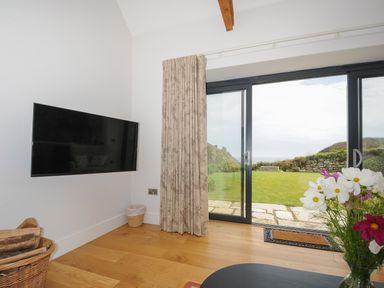 A room with a wall-mounted TV a basket of firewood patterned curtains sliding glass doors leading to a garden and flowers on a table at Pendower Cottage in Porthgwarra
