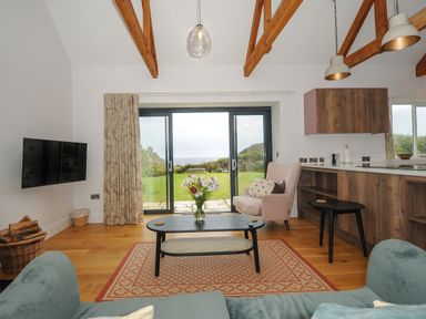 A living room with wooden beams on the ceiling sliding glass doors a television a coffee table with flowers a pink armchair and wooden cabinetry at Pendower Cottage in Porthgwarra