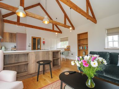 An open living room and kitchen with wooden beams a grey sofa a black coffee table with flowers and wooden shelves at Pendower Cottage in Porthgwarra