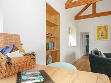 A dining area with a wicker basket and notebooks on the table an open wooden bookshelf with books a window a green chair a dark sofa exposed wooden ceiling beams and a framed painting on the wall at Pendower Cottage in Porthgwarra
