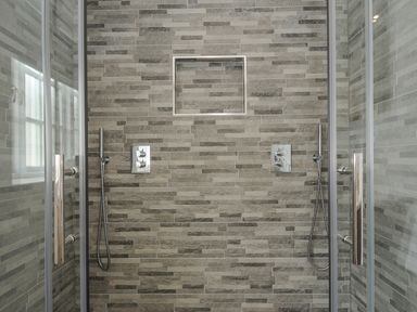 A shower area with two handheld showerheads and grey tiled walls at Pendower Cottage in Porthgwarra