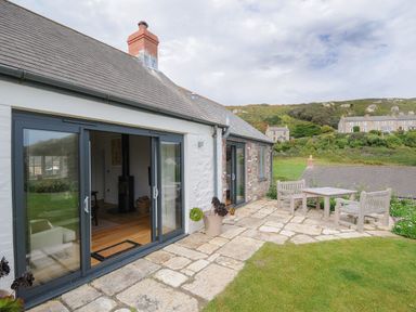 A stone patio with wooden outdoor furniture and sliding glass doors leading to a room at Pendower Cottage in Porthgwarra