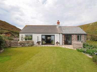 A cottage with two sliding glass doors a stone wall and a lawn surrounded by hills at Pendower Cottage in Porthgwarra
