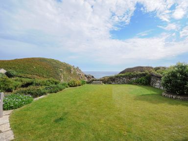 A grassy garden with stone walls and green shrubs overlooking hills and the sea at Pendower Cottage in Porthgwarra