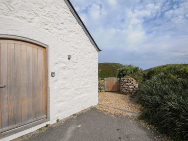 A white stone building with a wooden door next to a gravel path and wooden gate surrounded by bushes at Pendower Cottage in Porthgwarra