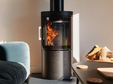 A modern wood-burning stove with fire inside next to a blue couch and a basket of firewood at Pendower Cottage in Porthgwarra