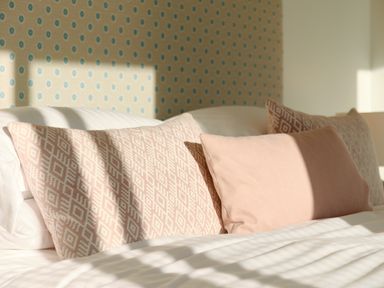 A bed with three pink patterned and plain cushions against a wall with blue dotted wallpaper at Pendower Cottage in Porthgwarra