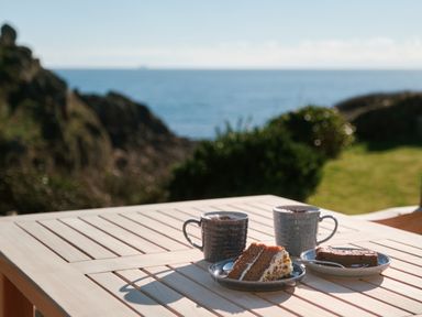 A wooden table outdoors with two mugs and plates of cake overlooking the sea at Pendower Cottage in Porthgwarra