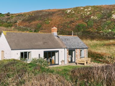 A cottage with white and stone walls surrounded by a grassy hillside with outdoor wooden furniture at Pendower Cottage in Porthgwarra