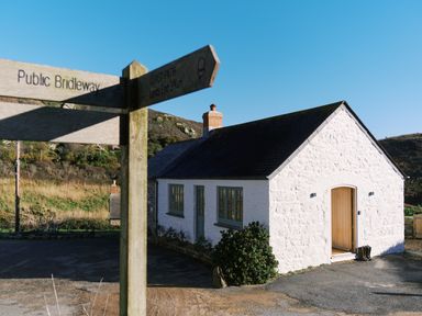 A white stone cottage with green windows and wooden door near a wooden signpost showing public bridleway and coast path directions at Pendower Cottage in Porthgwarra