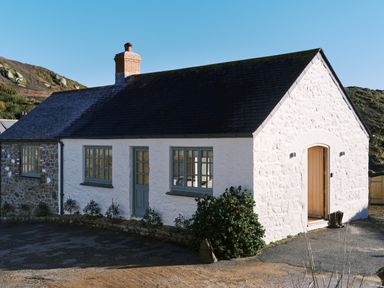 A white cottage with green doors and windows and a black roof surrounded by hills at Pendower Cottage in Porthgwarra