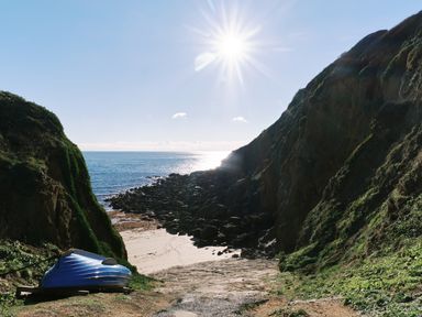 A rocky beach inlet with a blue boat on the shore and sun shining over the sea at Pendower Cottage in Porthgwarra