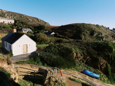A white stone cottage near hills with a path and a blue boat at Pendower Cottage in Porthgwarra