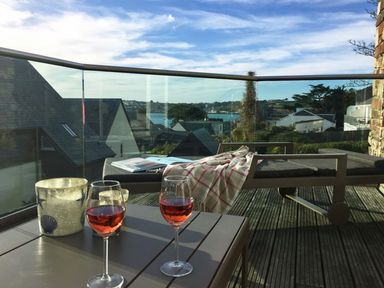 A balcony with two glasses of rose wine on a table and a lounge chair with a blanket overlooking rooftops and water at Gull Rock 4 in Rock