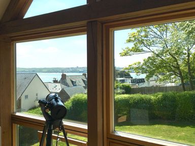 A telescope positioned inside a room with large windows overlooking rooftops a garden trees and a body of water at Gull Rock 4 in Rock