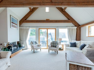 A living room with white sofas, armchairs, a wooden coffee table, large windows, exposed wooden beams, and a fireplace at Gull Rock 4 in Rock