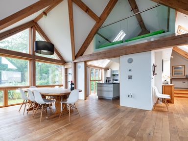 A dining area with a round wooden table and white chairs next to large windows in a room with wooden beams and a kitchen in the background at Gull Rock 4 in Rock