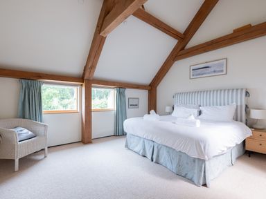 A bedroom with a large bed white linens a wicker chair two windows with blue curtains and wooden beams visible at Gull Rock 4 in Rock
