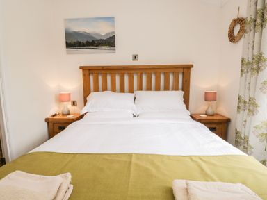 A bedroom with a wooden bed frame two pillows white bedding green throw two bedside tables with lamps and a wall picture at Pippin Lodge in Allithwaite near Cartmel