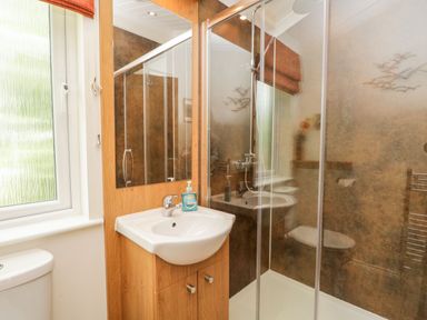 A bathroom with a sink and mirror next to a glass-enclosed shower and a frosted window at Pippin Lodge in Allithwaite near Cartmel