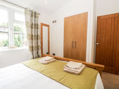 A bedroom with a bed featuring folded towels on a green runner a window with tree patterned curtains a wall mirror and wooden doors at Pippin Lodge in Allithwaite near Cartmel