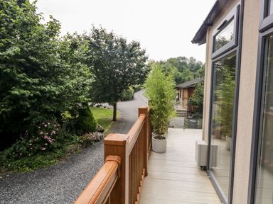 A wooden balcony with potted plants and glass doors overlooking a gravel path and trees at Pippin Lodge in Allithwaite near Cartmel