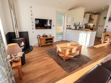 A living room with a wood burning stove wooden furniture and an open kitchen at Pippin Lodge in Allithwaite near Cartmel