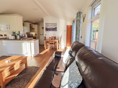 A living area with a brown leather sofa, wooden coffee table, dining table with chairs, and kitchen in the background at Pippin Lodge in Allithwaite near Cartmel
