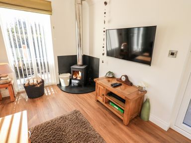 A corner of a living room with a wood burning stove a wall mounted television and a wooden cabinet at Pippin Lodge in Allithwaite near Cartmel