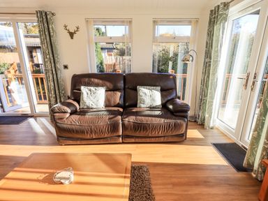 A living room with a brown leather sofa and cushions near windows and glass doors at Pippin Lodge in Allithwaite near Cartmel