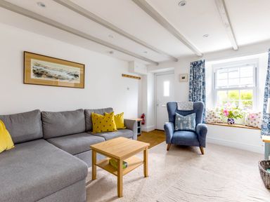 A living room with a gray sofa yellow pillows a blue armchair a wooden coffee table and a window seat with cushions and flowers at Stokes Cottage in St Minver