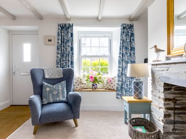 A living room with a blue armchair a window seat with cushions and flowers a side table with lamp and a stone fireplace at Stokes Cottage in St Minver