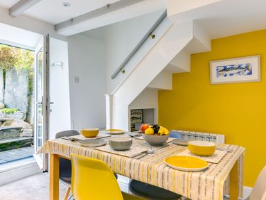 A dining area with a table set with bowls plates and cutlery and chairs underneath a staircase with a yellow wall and a framed fish picture at Stokes Cottage in St Minver