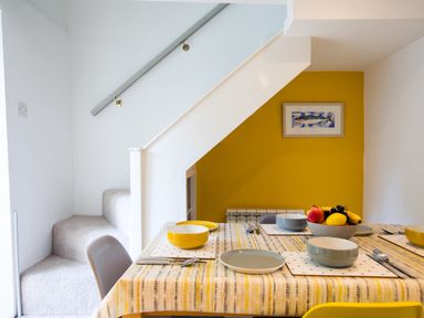 A dining table set with bowls plates and cutlery below a staircase with a yellow accent wall and a fish picture at Stokes Cottage in St Minver