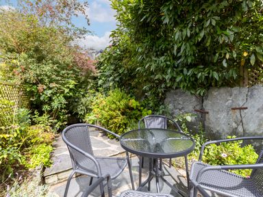 A glass top table with four black chairs on a stone patio surrounded by green plants and trees at Stokes Cottage in St Minver