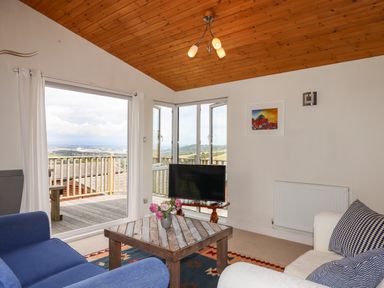 A living room with a wooden ceiling a coffee table with flowers a blue armchair a white sofa a television and large windows with a view at Tamar View Lodge Millbrook