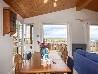 A dining room with a wooden table set with mugs and a vase of flowers next to a blue sofa overlooking a deck with countryside views at Tamar View Lodge Millbrook