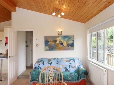 A living room with a chair and a sofa covered with a mermaid patterned blanket under a painting and near a window at Tamar View Lodge in Millbrook