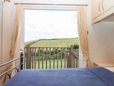 A bedroom with a bed covered in a blue blanket facing a window with curtains overlooking a wooden fence and hillside at Tamar View Lodge in Millbrook