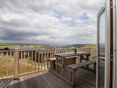 A wooden deck with a picnic table and benches overlooking countryside and wooden cabins at Tamar View Lodge in Millbrook