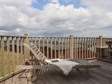 A wooden deck chair with a blanket and book on a wooden deck with a wooden railing and houses in the background at Tamar View Lodge in Millbrook