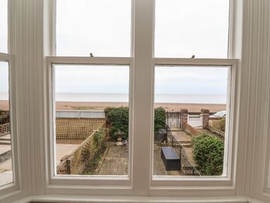 A view through three white framed windows showing a patio garden with bushes and a gate opening to a beach at 42 Crag Path in Aldeburgh