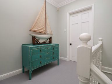 A hallway with a teal chest of drawers and a model sailboat on top next to a white door and white staircase railing at 42 Crag Path in Aldeburgh