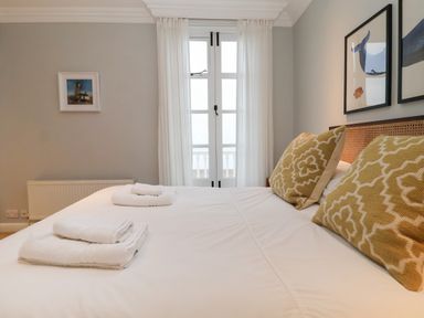 A bedroom with a bed having two patterned pillows white towels on a bed and a window with white curtains at 42 Crag Path in Aldeburgh