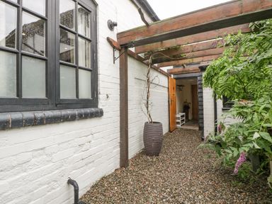 A gravel pathway with a large planter under a wooden pergola beside a white brick wall with black framed windows at The Old Bakehouse in Pembridge