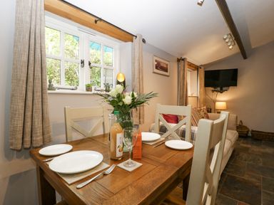 A dining area with a wooden table set with plates and cutlery a vase of flowers and drinks next to a window and a living space with a sofa and a wall-mounted TV at The Old Bakehouse in Pembridge